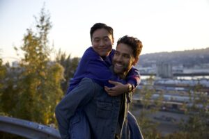 Photo of two smiling people outside at sunset, one person giving the other a piggyback ride. The city of Portland can be seen out of focus in the distance. 