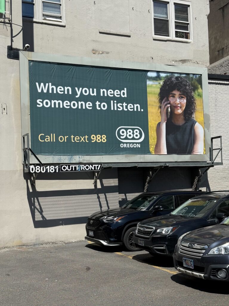 Outdoor billboard in a parking lot next to cars featuring a message from 988. Headline reads, "When you need someone to listen." Subhead reads "call or text 988." Includes the 988 Oregon logo and a image of a young woman with curly brown hair in a black tank top holding a cell phone to their ear and looking directly into the camera.