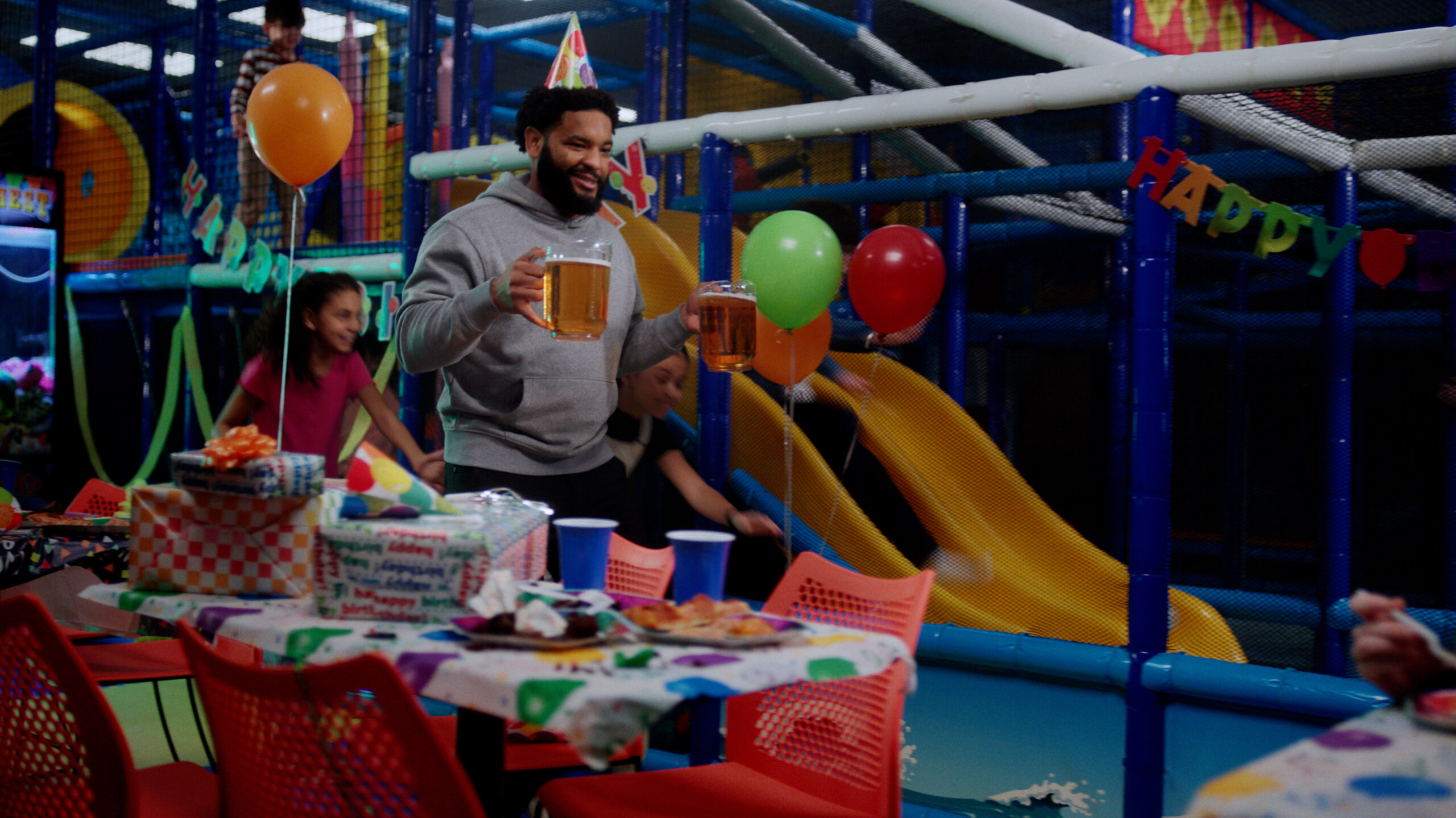 A man wearing a birthday party hat carries two large mugs of beer while walking through a children’s indoor playground decorated for a party. Kids run and play around him near colorful slides, balloons, and a table covered with presents, cups, and snacks.