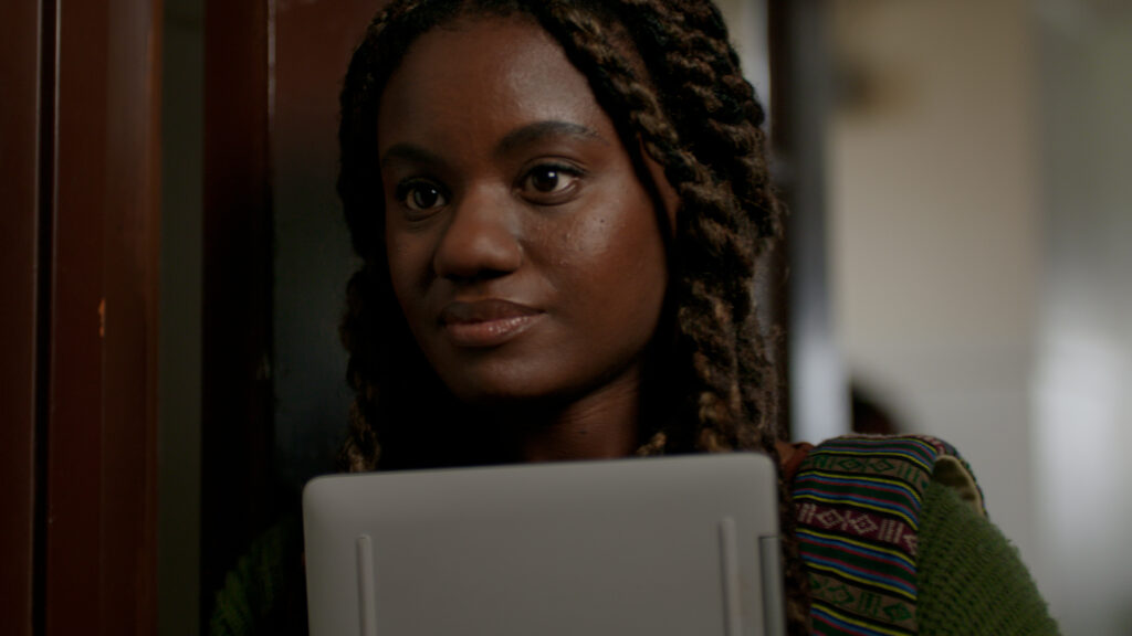 close up photo of a young, black teenager girl standing in a school hallway holding a laptop close to their body.