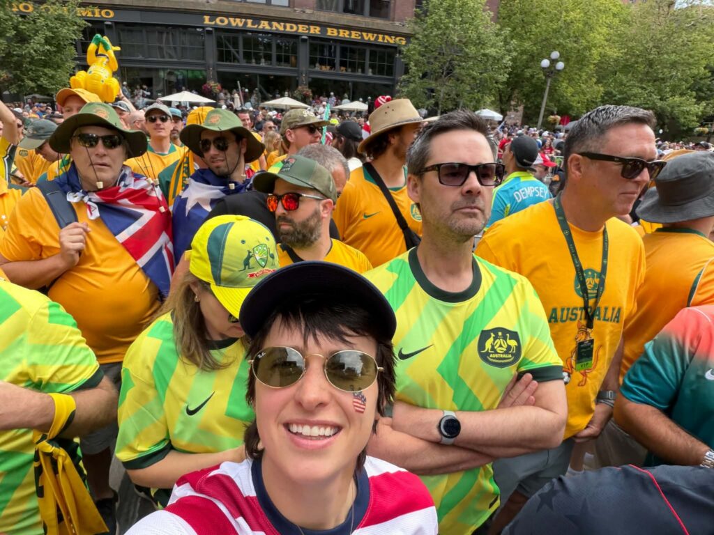 Kelsey Overbey, a team USA soccer fan wearing a U.S. jersey and sunglasses smiles for a selfie while standing among a large crowd of Australian soccer fans gathered in downtown Seattle before a match.
